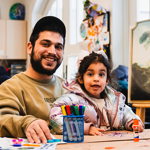 A man and young girl sit at a table with markers and art supplies