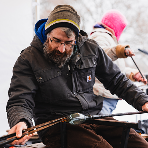 A man shapes a clear glass piece with tongs
