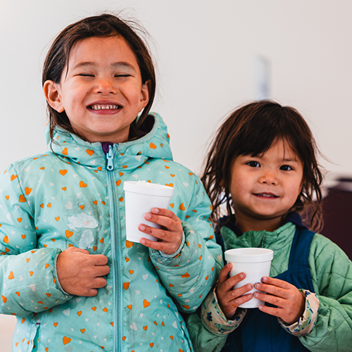 Two young girls in winter coats smile while holding cups of hot chocolate