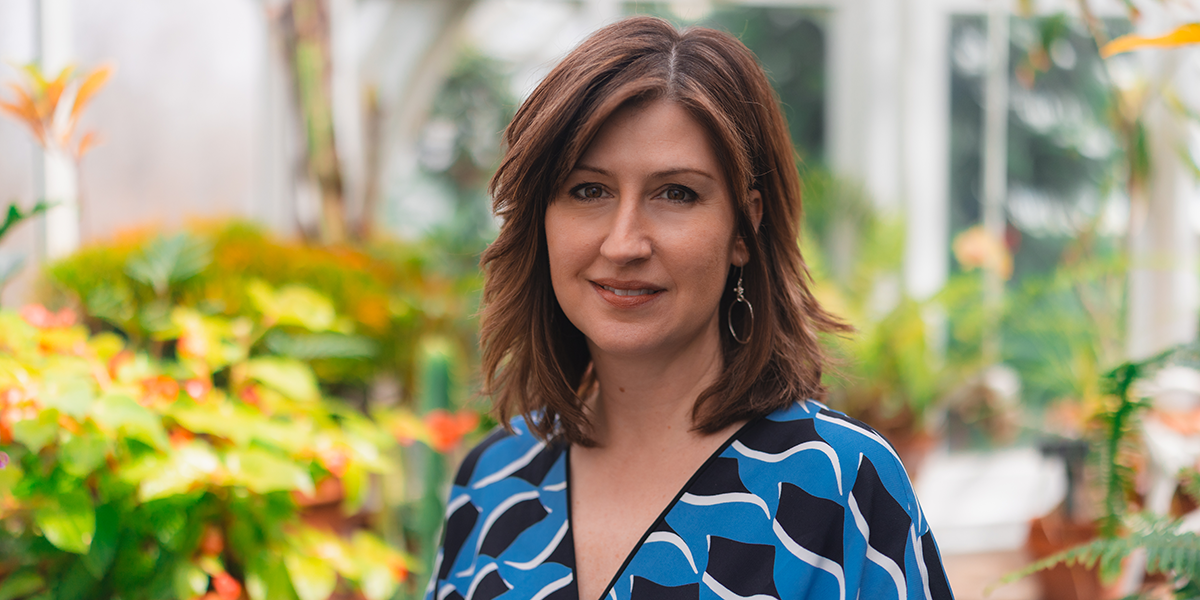 A woman with medium length brown hair wears a blue and black patterned dress and stands in a greenhouse